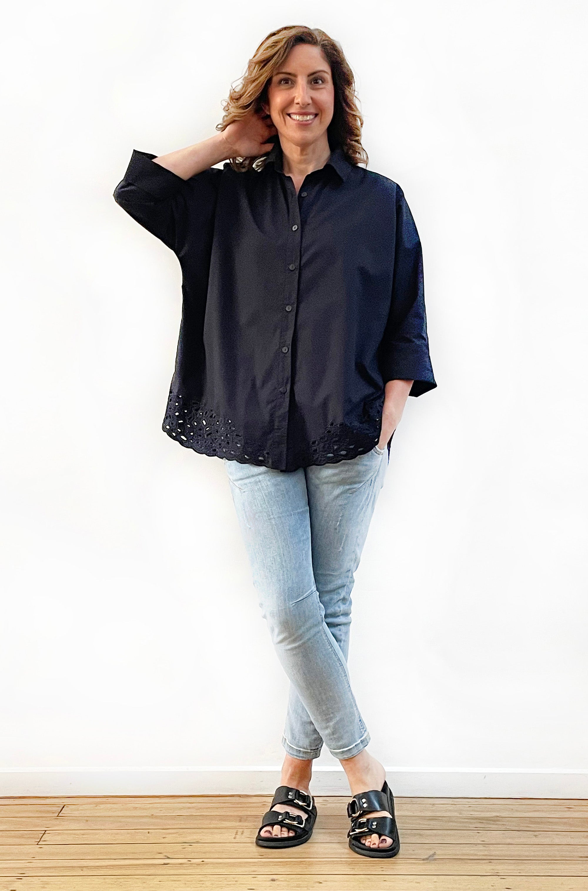 A woman standing on a wooden floor wearing a black short-sleeved shirt with kimono sleeves and button-through design, paired with blue jeans and black flat sandals.